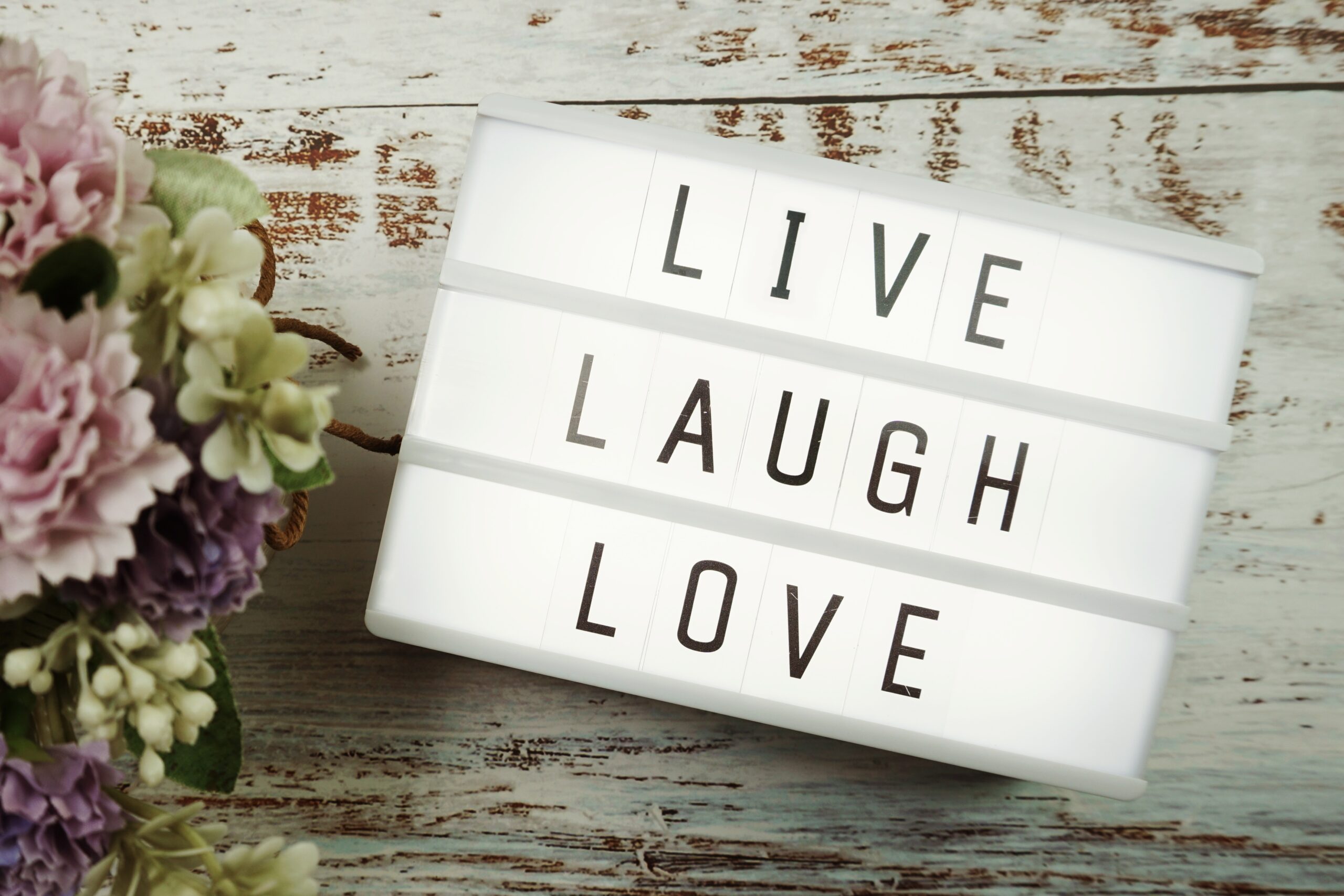 Picture of a white Live Laugh Sign on a white table, on the left there are flowers